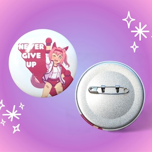 May include: Two round buttons. One button features an anime-style character with pink hair and the text "NEVER GIVE UP". The other button is silver with a safety pin on the back. The background is a gradient of pink and purple.