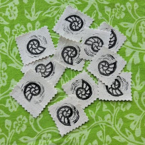 May include: A collection of small, square, off-white fabric patches with pinked edges, each featuring a black ink-stamped ammonite fossil design. The patches are arranged in a scattered cluster, displaying a consistent graphic print of a spiral shell.