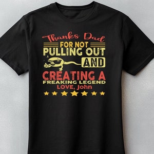 Puede incluir: Camiseta negra con el texto "Gracias papá por no retirarte y crear una leyenda, Love, John." El diseño incluye un espermatozoide con gafas de sol. El texto es rojo y amarillo.