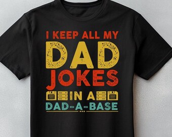 May include: Black t-shirt with the text "I KEEP ALL MY DAD JOKES IN A DAD-A-BASE" in yellow, orange, and teal. The design includes a retro computer floppy disk graphic. A fun, novelty tee.