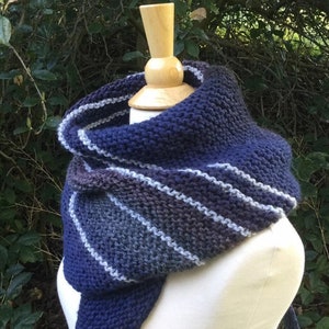 K&ouml;nnte beinhalten: Ein gestrickter Schal in Marineblau, Grau und Wei&szlig;, pr&auml;sentiert auf einer Schaufensterpuppe. Der Schal hat ein Streifenmuster und eine dreieckige Form, ideal, um W&auml;rme und Stil zu verleihen. Der Hintergrund zeigt gr&uuml;nes Laub.