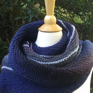 K&ouml;nnte beinhalten: Ein handgestrickter Schal in Blau-, Grau- und Schwarzt&ouml;nen, auf einer Schaufensterpuppe pr&auml;sentiert. Der Schal hat ein strukturiertes Strickmuster und einen Farbverlauf, der von Dunkelblau zu helleren Farbt&ouml;nen &uuml;bergeht.