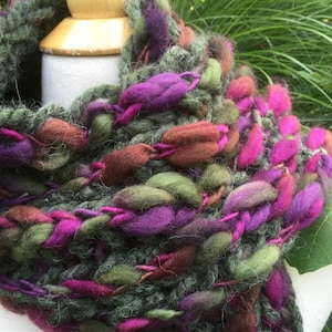 May include: A chunky knit scarf featuring a blend of thick, textured yarn in shades of forest green, deep purple, magenta, and earthy brown. The yarn is woven in a loose, open stitch pattern, creating a voluminous and tactile appearance throughout.