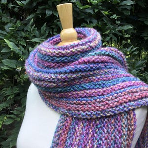 Op de afbeelding: Handgebreide sjaal in paarse, blauwe en roze tinten, getoond op een witte mannequin. De sjaal heeft een gestreept patroon en een zachte textuur, perfect voor de winter.