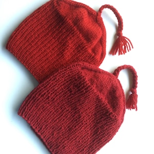 May include: Two bright red knitted hats, each featuring a ribbed texture and a braided yarn cord attached to the top, ending in a small tassel. The hats are crafted from a solid red yarn and display a consistent knit pattern throughout the design.