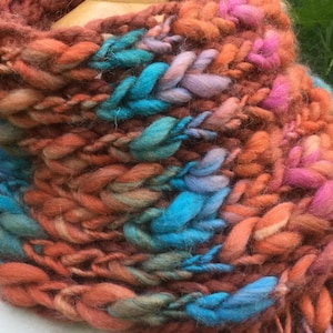 May include: A chunky knit scarf featuring a thick, braided texture. The yarn is a variegated blend of burnt orange, deep rust, bright turquoise, soft lavender, and vibrant pink, creating a multi-colored, textured appearance throughout the entire piece.