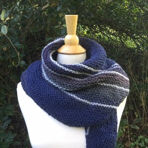 K&ouml;nnte beinhalten: Ein gestrickter Schal in Marineblau, Grau und Wei&szlig;, der auf einer Schaufensterpuppe pr&auml;sentiert wird. Der Schal hat horizontale Streifen und ein dickes, strukturiertes Strickmuster. Der Hintergrund ist eine verschwommene Ansicht von gr&uuml;nem Laub.