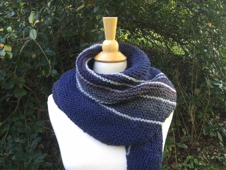 K&ouml;nnte beinhalten: Ein gestrickter Schal in Marineblau, Grau und Wei&szlig;, der auf einer Schaufensterpuppe pr&auml;sentiert wird. Der Schal hat horizontale Streifen und ein dickes, strukturiertes Strickmuster. Der Hintergrund ist eine verschwommene Ansicht von gr&uuml;nem Laub.