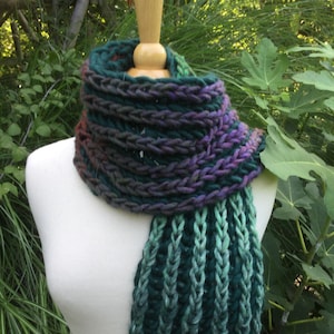 May include: A chunky knit scarf featuring a variegated color palette of deep forest green, teal, and dark purple. The scarf is wrapped in a circular cowl style around the neck, with a long, vertically ribbed tail hanging down in shades of teal.
