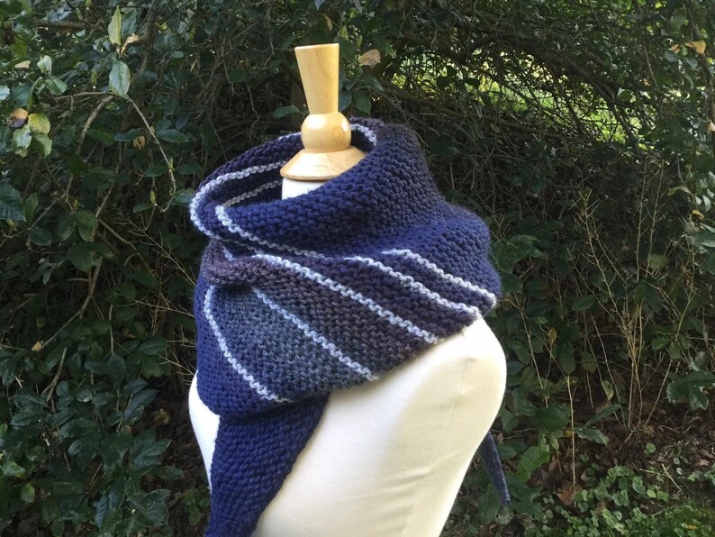 K&ouml;nnte beinhalten: Ein gestrickter Schal in Marineblau, Grau und Wei&szlig;, pr&auml;sentiert auf einer Schaufensterpuppe. Der Schal hat ein Streifenmuster und eine dreieckige Form, ideal, um W&auml;rme und Stil zu verleihen. Der Hintergrund zeigt gr&uuml;nes Laub.