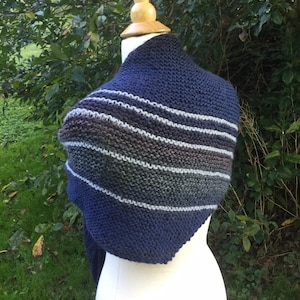 K&ouml;nnte beinhalten: Ein marineblauer Strickschal mit horizontalen Streifen in Grau-, Braun- und Wei&szlig;t&ouml;nen. Der Schal wird auf einer wei&szlig;en Schaufensterpuppe vor einem Hintergrund aus gr&uuml;nem Laub gezeigt. Die Strickstruktur ist deutlich sichtbar.