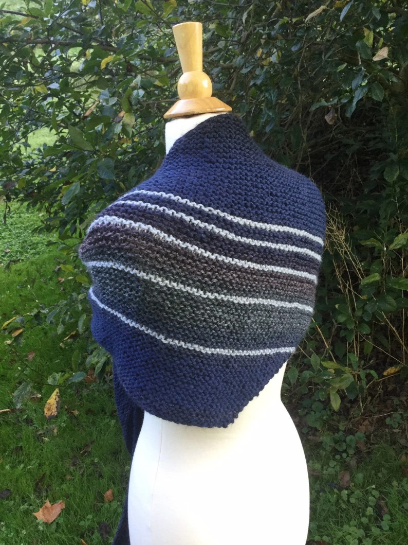K&ouml;nnte beinhalten: Ein marineblauer Strickschal mit horizontalen Streifen in Grau-, Braun- und Wei&szlig;t&ouml;nen. Der Schal wird auf einer wei&szlig;en Schaufensterpuppe vor einem Hintergrund aus gr&uuml;nem Laub gezeigt. Die Strickstruktur ist deutlich sichtbar.