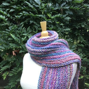 Op de afbeelding: Een handgebreide sjaal in tinten paars, blauw en roze is over een mannequin gedrapeerd. De sjaal is gemaakt van dik garen, met een getextureerd, grof gebreid patroon. De achtergrond heeft weelderig groen gebladerte.