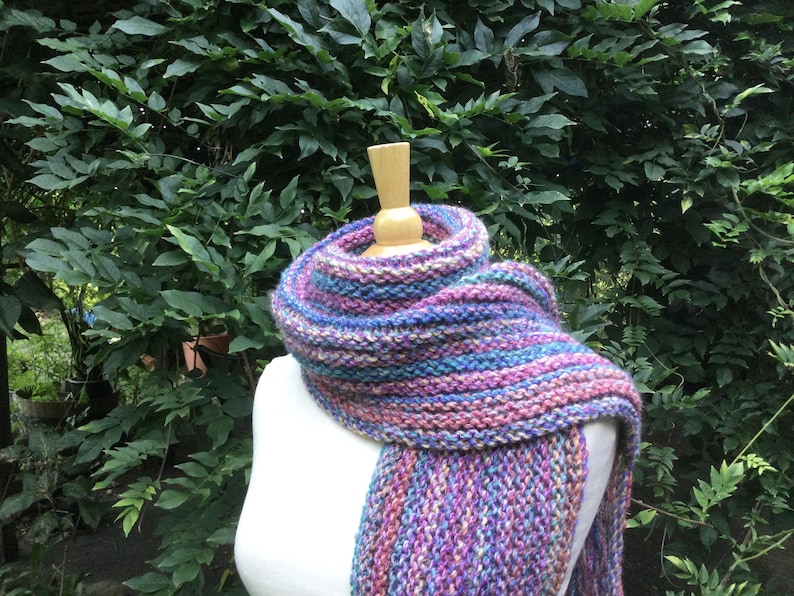 Op de afbeelding: Een handgebreide sjaal in tinten paars, blauw en roze is over een mannequin gedrapeerd. De sjaal is gemaakt van dik garen, met een getextureerd, grof gebreid patroon. De achtergrond heeft weelderig groen gebladerte.