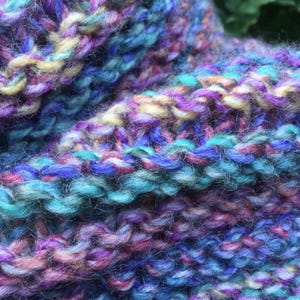Op de afbeelding: Close-up van een handgebreid item met een textuurrijk, kleurrijk patroon. Het garen heeft een mix van blauw, paars, roze en geel. De gebreide stof heeft een zachte, dikke uitstraling, wat wijst op een gezellig accessoire zoals een sjaal of col.