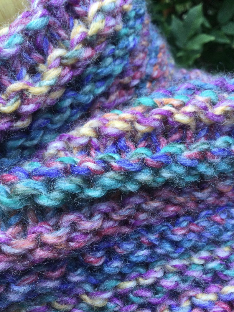 Op de afbeelding: Close-up van een handgebreid item met een textuurrijk, kleurrijk patroon. Het garen heeft een mix van blauw, paars, roze en geel. De gebreide stof heeft een zachte, dikke uitstraling, wat wijst op een gezellig accessoire zoals een sjaal of col.
