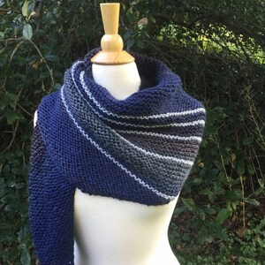 K&ouml;nnte beinhalten: Ein handgestrickter Schal in Marineblau, Grau und Wei&szlig;, der auf einer Schaufensterpuppe pr&auml;sentiert wird. Der Schal hat ein Streifenmuster und einen langen, drapierten &Auml;rmel. Der Hintergrund ist gr&uuml;nes Laub.
