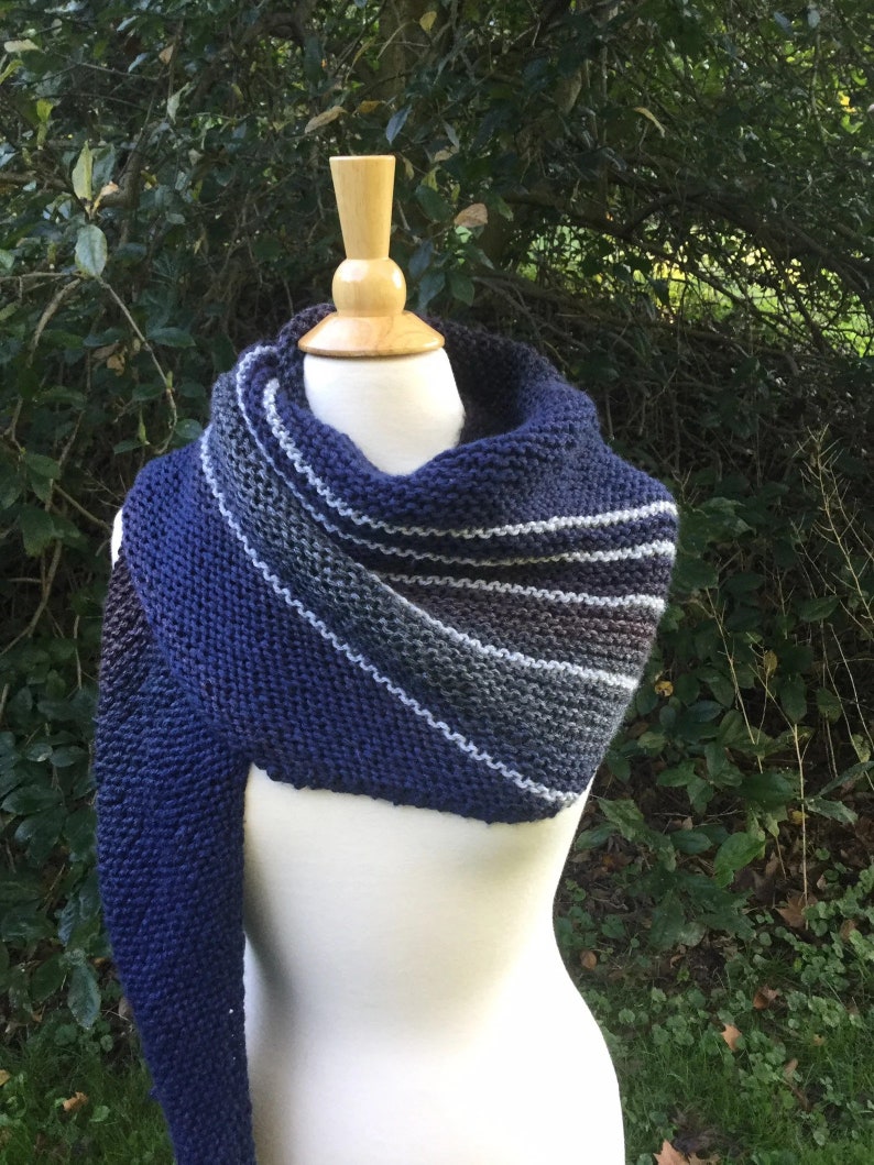 K&ouml;nnte beinhalten: Ein handgestrickter Schal in Marineblau, Grau und Wei&szlig;, der auf einer Schaufensterpuppe pr&auml;sentiert wird. Der Schal hat ein Streifenmuster und einen langen, drapierten &Auml;rmel. Der Hintergrund ist gr&uuml;nes Laub.