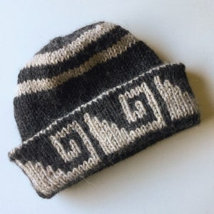 May include: A dark brown and beige knit beanie hat. The hat features horizontal stripes on the crown and a geometric pattern around the brim. The hat is made of a soft, textured yarn and is folded at the brim.