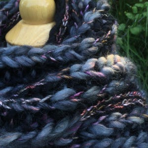 May include: A chunky knit cowl scarf featuring thick, braided yarn in shades of dark blue, black, and grey, with interwoven metallic copper and purple threads. The textured, heavy-weight yarn is wrapped in multiple layers around a wooden display form.