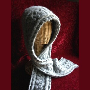 May include: A light gray knitted hood and scarf set. The hood is designed with a cable knit pattern and a decorative edge. The scarf is long and features the same cable knit design. The set is displayed on a wicker mannequin head against a red background.