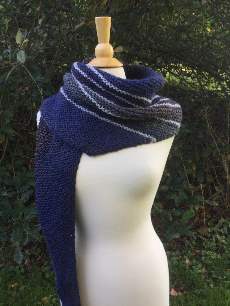 May include: A hand-knitted scarf in shades of navy blue, gray, and white is draped over a mannequin. The scarf features a cowl neck and a long, rectangular section. The color gradient transitions from dark blue to gray and white stripes.