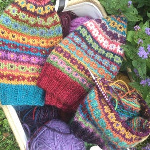 May include: A collection of colorful, hand-knitted hats and yarn in a basket. The hats feature intricate patterns in shades of blue, red, orange, and green. A pair of knitting needles are in use, with a partially completed hat.