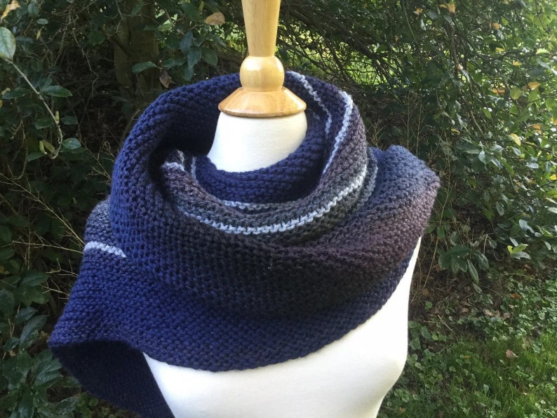 K&ouml;nnte beinhalten: Ein handgestrickter Schal in Blau-, Grau- und Schwarzt&ouml;nen, auf einer Schaufensterpuppe pr&auml;sentiert. Der Schal hat ein strukturiertes Strickmuster und einen Farbverlauf, der von Dunkelblau zu helleren Farbt&ouml;nen &uuml;bergeht.