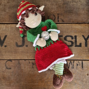 May include: A crocheted elf doll wearing a green and red outfit with a white trim. The elf is holding a silver disco ball and is lying on a wooden surface.