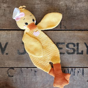 May include: A handmade yellow crochet duckling with orange feet and beak, wearing a white bonnet with a pink bow. The duckling has black eyes and a pink heart on its chest. The item is lying on a wooden surface.