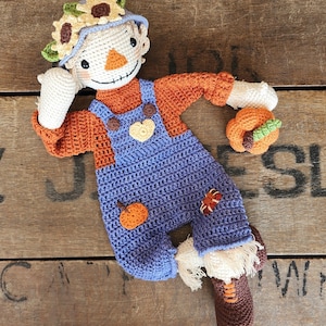 Op de afbeelding: Een gehaakte vogelverschrikkerpop met een zonnebloemhoed, oranje en blauwe overalls en een kleine pompoen in zijn hand.