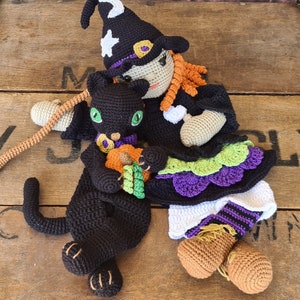 May include: A black crocheted cat with a pumpkin in its paws sits next to a crocheted witch doll wearing a black and purple dress with a green and white trim.
