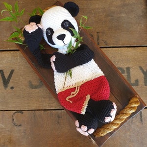Könnte beinhalten: Ein gehäkelter Pandabär, der rote und weiße Shorts trägt. Der Panda liegt auf einer Holzoberfläche mit einem daran befestigten Seil.