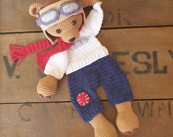 Puede incluir: Un oso de peluche de crochet marrón que lleva un suéter blanco, pantalones azules con un parche circular rojo y un gorro de aviador de cuero marrón con gafas. El oso está acostado sobre una superficie de madera.