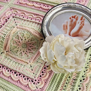 May include: A handmade crochet blanket in pastel pink, green, and cream tones. A silver oval picture frame with a photo of baby feet and a large white peony flower are on the blanket.