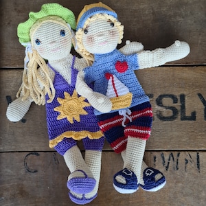 May include: Two crocheted dolls, one wearing a purple and yellow dress and a green hat, the other wearing a blue and red outfit with a white hat. Both dolls have yarn hair and are smiling.