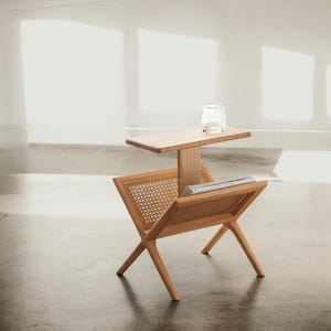 May include: A wooden side table with a magazine rack and a small top shelf. The table is made of natural wood and has a woven rattan insert. The table is in a light-colored room with a white wall in the background.