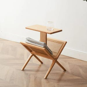 May include: A wooden side table with a woven rattan magazine rack. The table has a small upper shelf and a lower shelf for magazines. The table is made of natural wood and has a light brown finish.