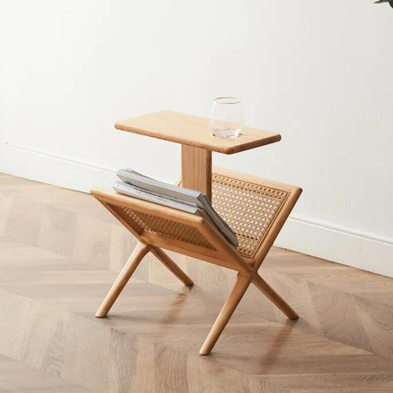 May include: A wooden side table with a woven rattan magazine rack. The table has a small upper shelf and a lower shelf for magazines. The table is made of natural wood and has a light brown finish.