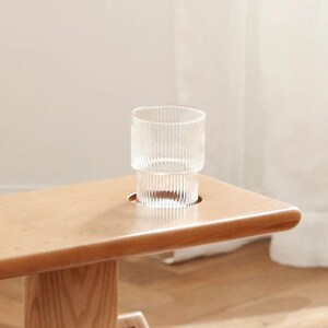 May include: A light wood table with a glass cup sitting in a cutout on the surface. The glass cup has a ribbed design.