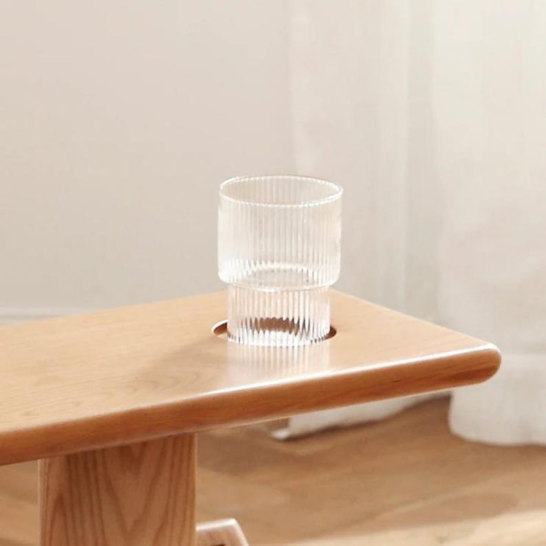May include: A light wood table with a glass cup sitting in a cutout on the surface. The glass cup has a ribbed design.
