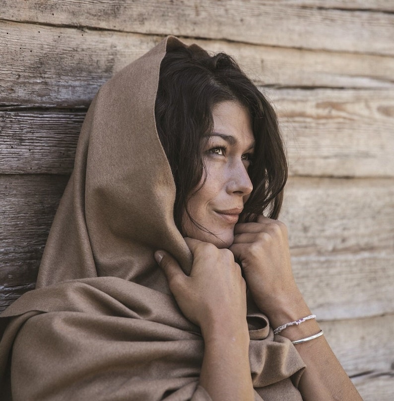 May include: A woman with long dark hair is wearing a brown hooded scarf. She is looking off to the side and smiling.