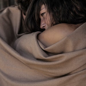 May include: A close-up of two people wrapped in a brown blanket. The person in the foreground is looking off to the side with a thoughtful expression.