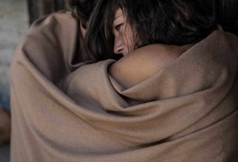 May include: A close-up of two people wrapped in a brown blanket. The person in the foreground is looking off to the side with a thoughtful expression.