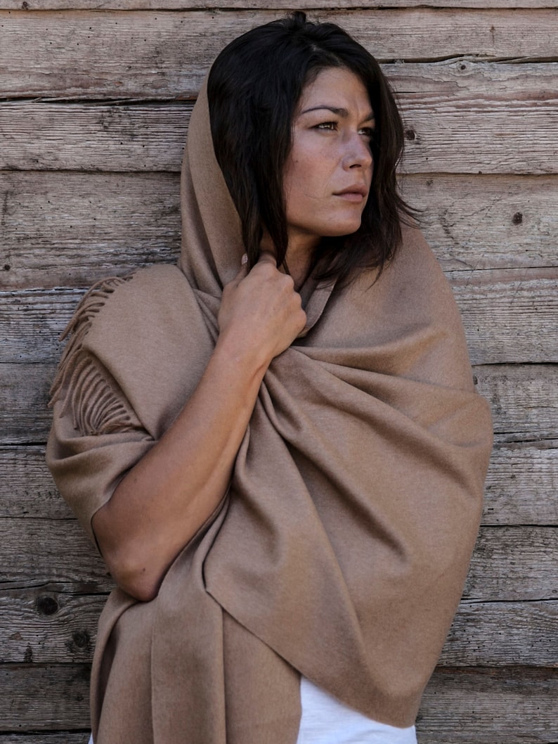 May include: A brown shawl with fringe detail is draped around a person's shoulders. The shawl is made of a soft, luxurious fabric and is perfect for keeping warm on a chilly day.