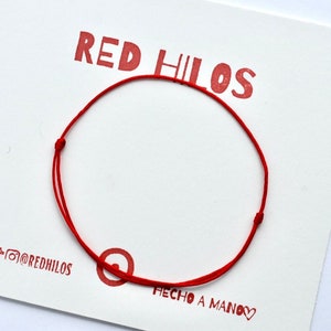 May include: A red string bracelet with a knot on each end. The bracelet is on a white card with the text "RED HILOS" and "HECHO A MANO" in red.