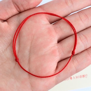 May include: A bright red string bracelet, knotted at the ends, is held in an open hand. The bracelet is circular, and the red cord is thin. The background is a light color, and a small card is visible.