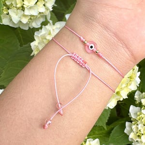 Puede incluir: Dos pulseras de cuerda rosa con un dije de ojo turco rosa en una de las pulseras. Las pulseras están atadas con un nudo y tienen pequeñas cuentas transparentes en los extremos.