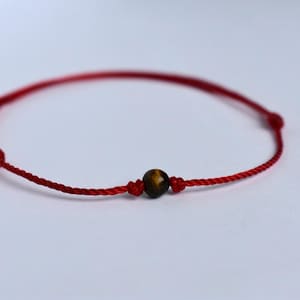May include: Red string bracelet with a central, round, tiger's eye bead. The bracelet is knotted and adjustable. The tiger's eye bead is brown with golden and black streaks. The bracelet is on a white background.