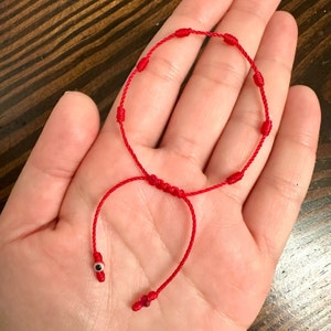 May include: Red string bracelet with a small evil eye charm. The bracelet is tied with knots and has a sliding knot for adjusting the size.